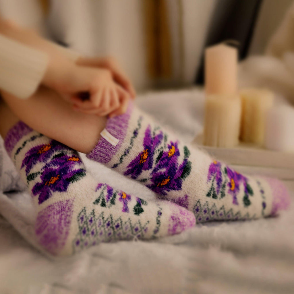 Women's legs on bed wearing goat hair socks with purple violets. 