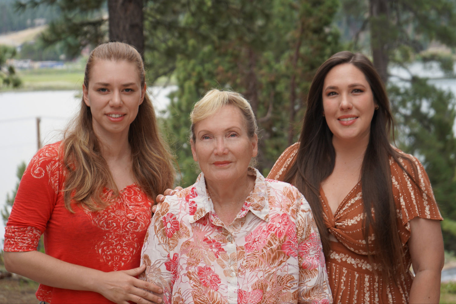 Three women standing together outdoors with a lake and trees in the background