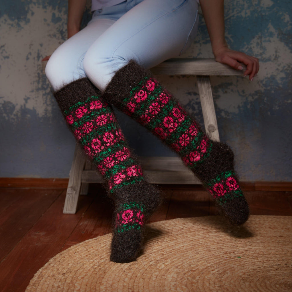 Woman’s legs sitting on a chair wearing black goat wool socks knee-high length with a pink and green flower design.