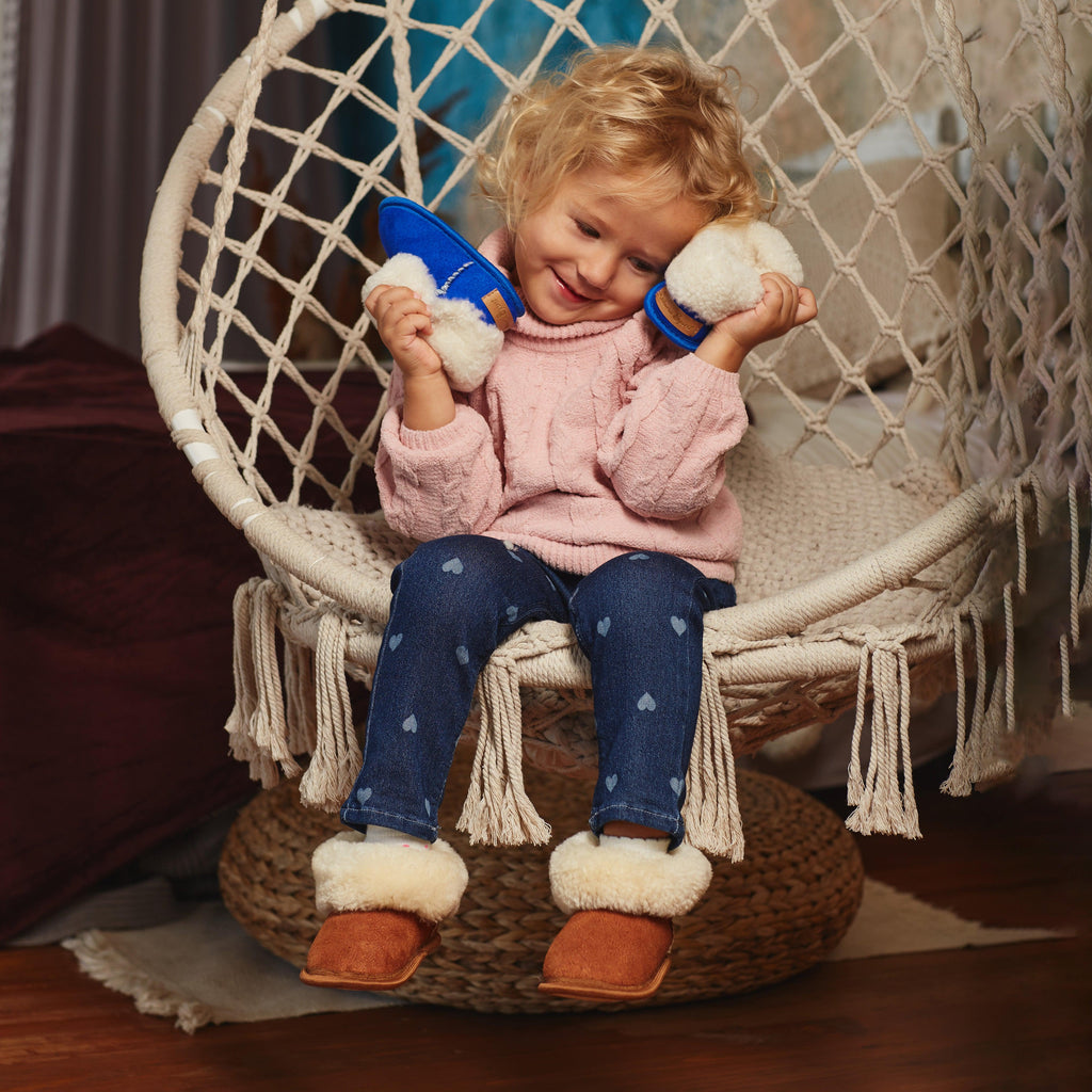 Child sitting in hanging chair wearing brown sheepskin slippers holding blue sheepskin slippers. 