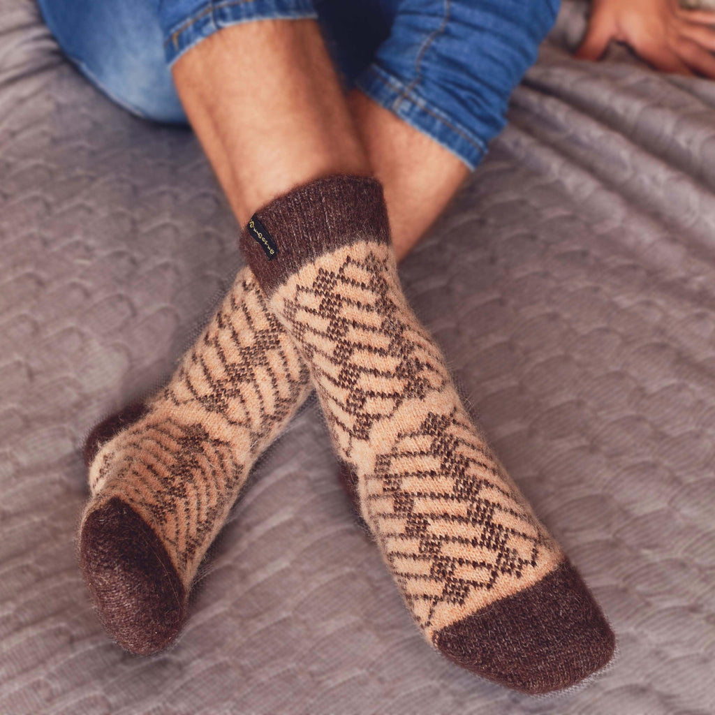 Man's legs on the bed wearing brown merino wool crew socks with geometric patterns.