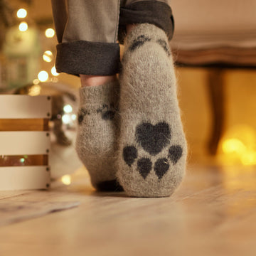 Person's feet in home interior wearing gray pants and gray low-cut goat wool warm socks with a black paw-print design.