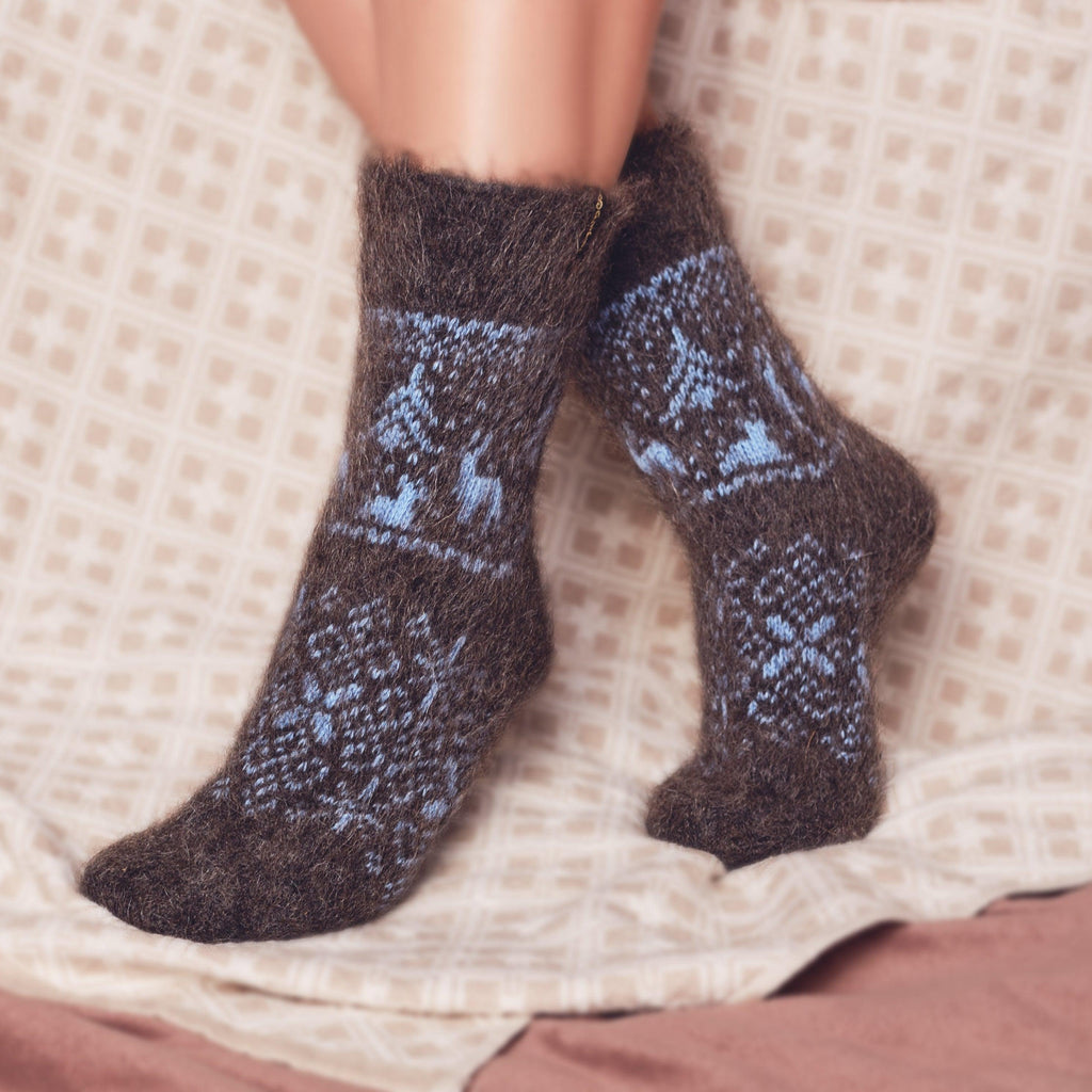 Woman’s legs near a bed wearing relaxed-fit goat wool charcoal crew socks with blue deer, trees, and winter ornaments.