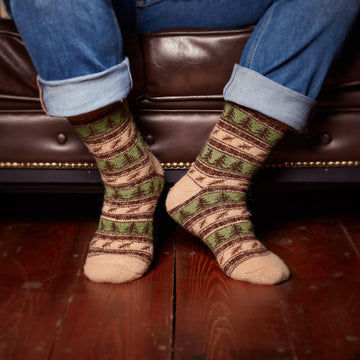 Man’s legs in home interior wearing jeans and brown green wool socks crew length with forest design.