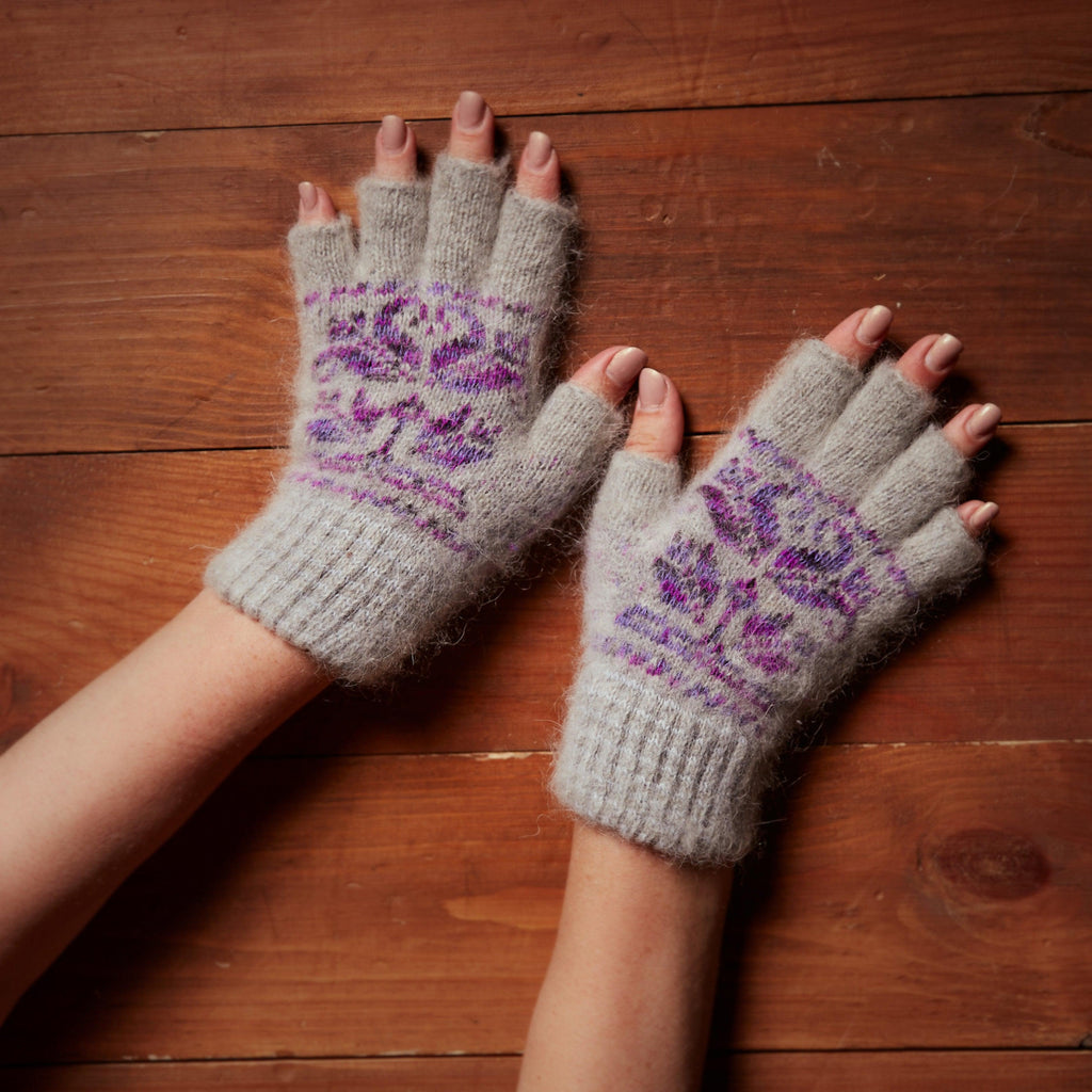 Person's hands wearing light-gray thick goat wool fingerless gloves with a purple stylized flower design.