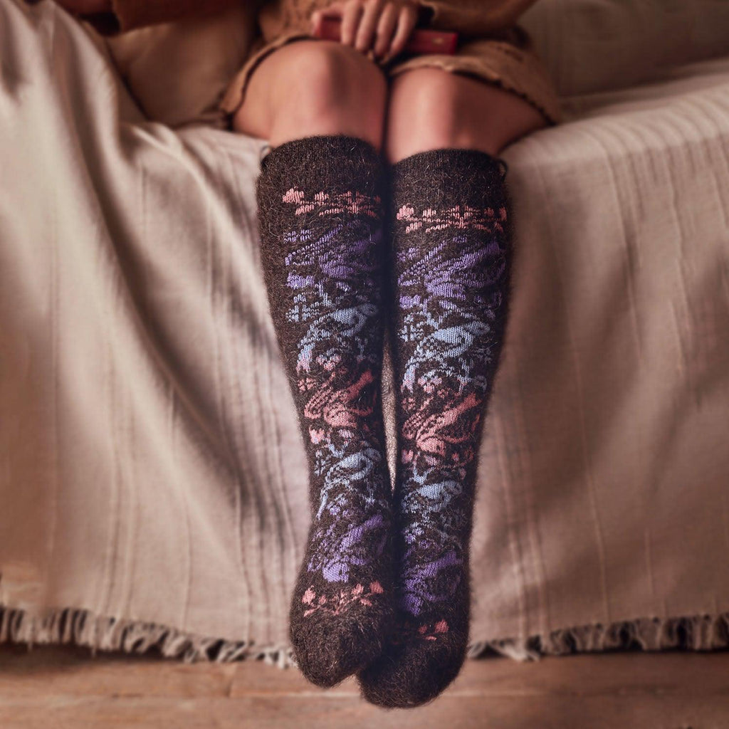 Woman's legs sitting on the bed wearing fuzzy warm goat hair black knee-high socks with pink, purple, and blue birds.