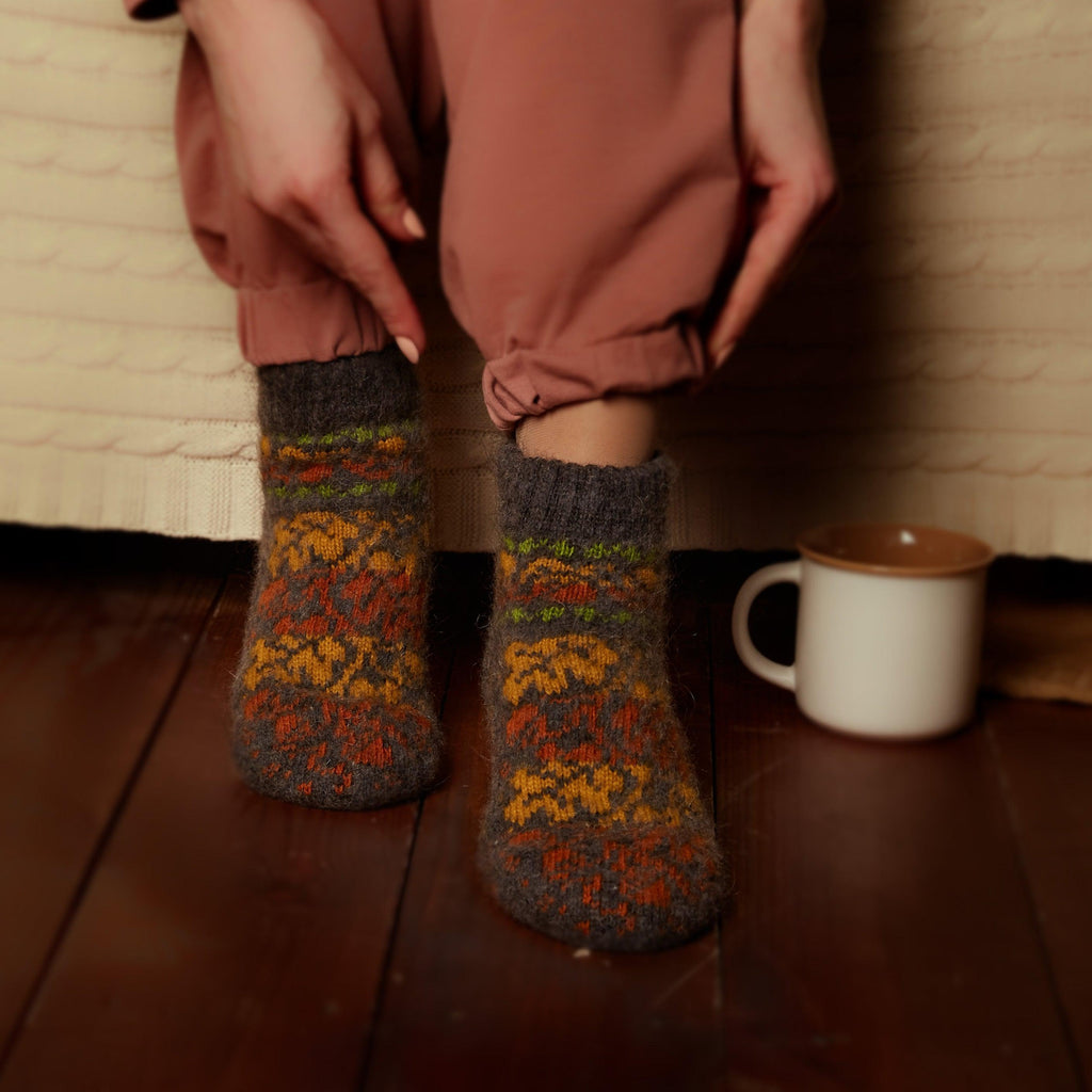 Woman’s legs sitting on the bed wearing pants and warm goat hair black low-cut socks with yellow and orange fall leaves.