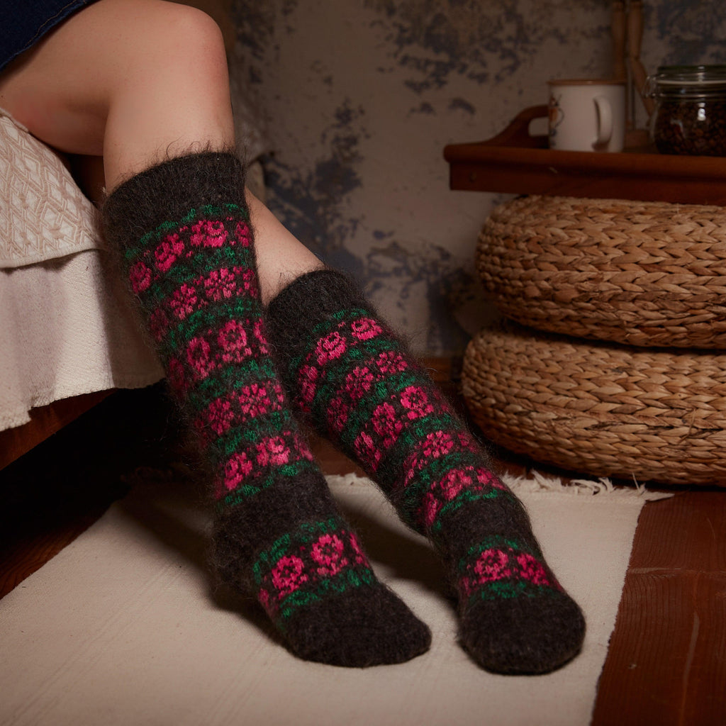 Woman's legs in home interior wearing black knee-high goat hair socks with green and bright pink flower design.
