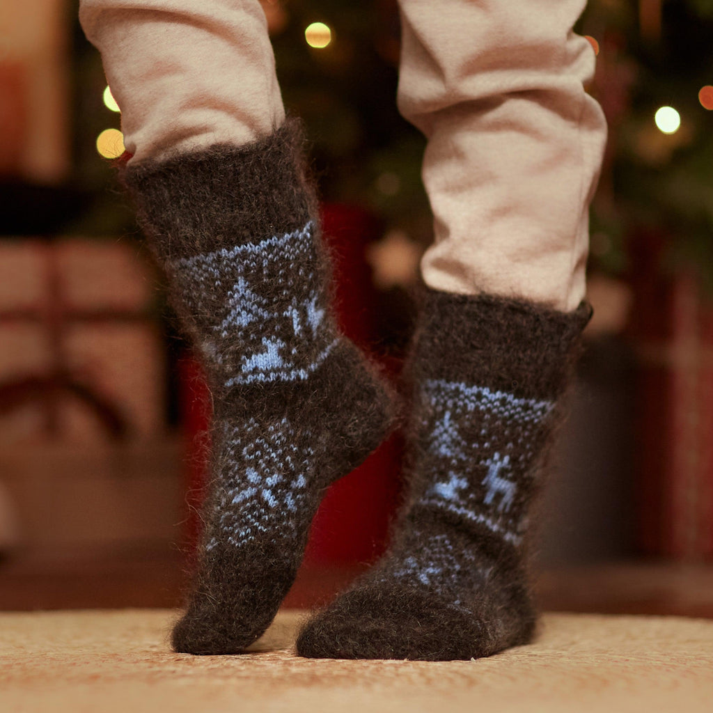 Person's legs in home interior wearing pants and crew-length no elastic goat hair charcoal socks with blue deer, trees, and winter ornaments.
