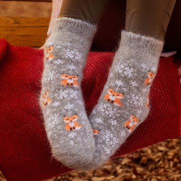 Woman's legs in outdoor settings wearing brown pants and grey goat wool crew socks with foxes and snowflakes.