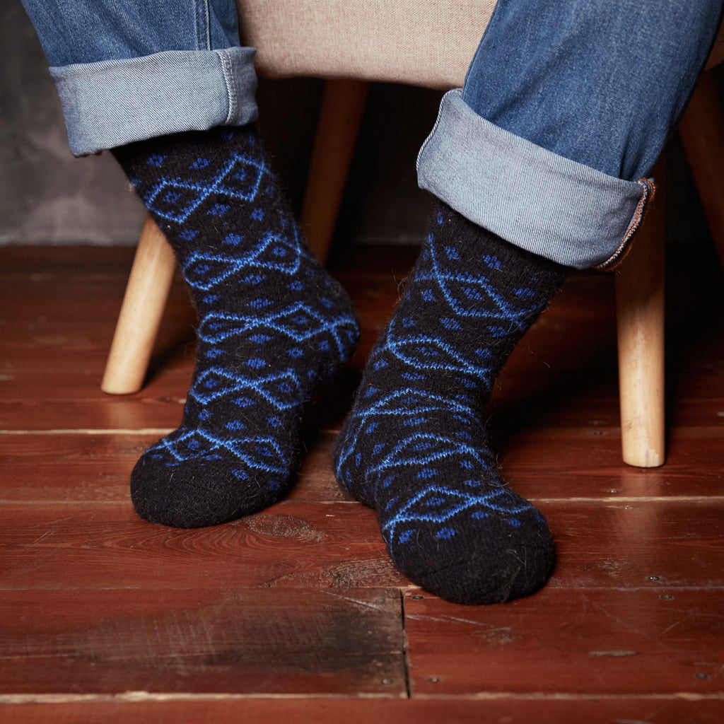 Man's legs in home interior wearing black crew-length merino wool socks with diamond-shaped blue design.