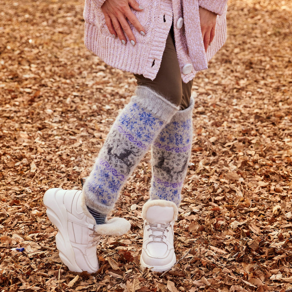 Woman's legs posing outdoors wearing grey goat hair leg warmers with blue snowflakes and black deer design.