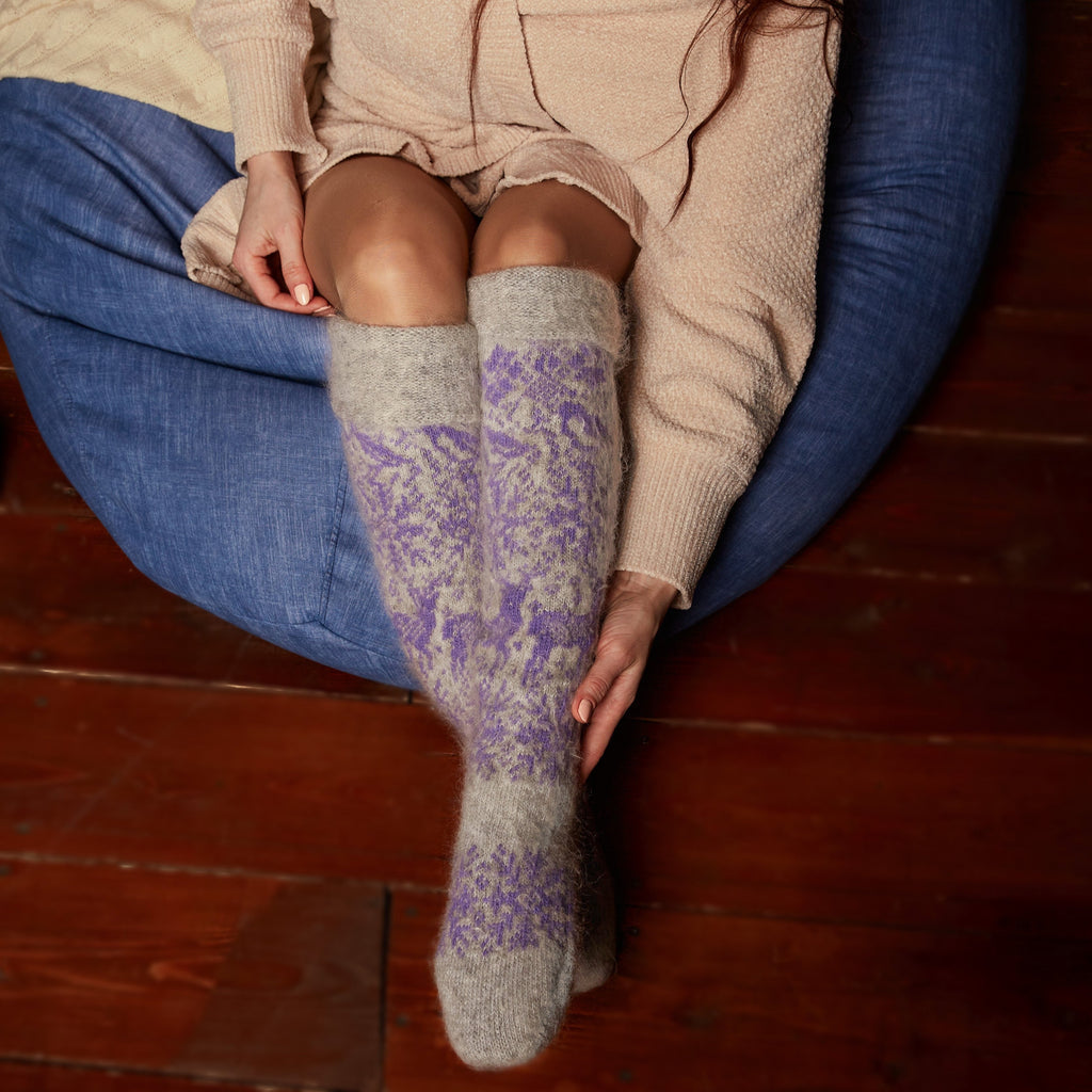 Woman's legs wearing grey over-the-knee goat wool socks with purple winter designs.
