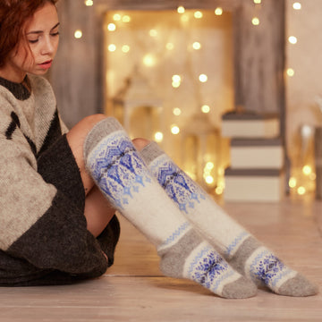 Woman sitting on the wooden floor wearing white goat hair and grey knee-high relaxed-fit socks with blue winter ornament.